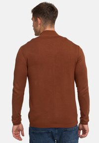 Brun strikket genser med lange ermer og ribbestrikket krage, som har en teksturert stoff og en liten logo på nederste del.