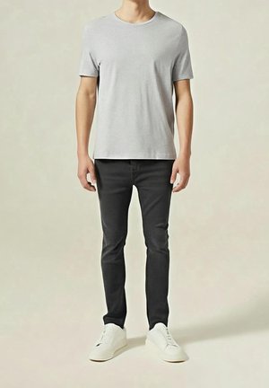 Man wearing light gray short-sleeve t-shirt, slim black pants, and white sneakers standing against plain neutral background.