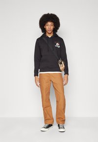 Fekete kapucnis pulóver grafikai designnal, barna nadrággal és fekete sportcipővel párosítva. Tartalmaz egy válltáskát és egy fehér aláöltözetet.