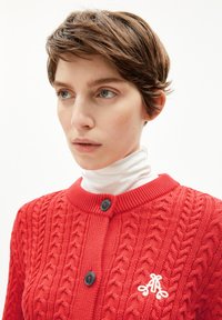 Roter gestrickter Cardigan mit einem strukturierten Kabelmuster, rundem Kragen, zwei schwarzen Knöpfen und einem weißen gestickten Logo auf der linken Brust.