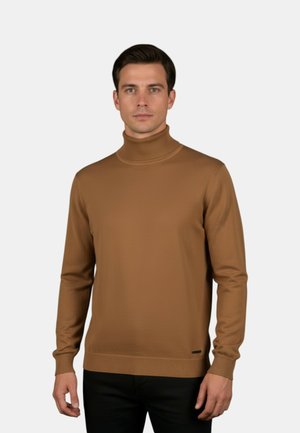 Uomo che indossa un maglione marrone a collo alto e pantaloni neri, in piedi davanti a uno sfondo grigio chiaro uniforme.