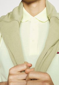 Béžový svetr přehozený přes světle žluté polo, s texturovanou tkaninou a dvěma knoflíky na límci. Ruce drží svetr.