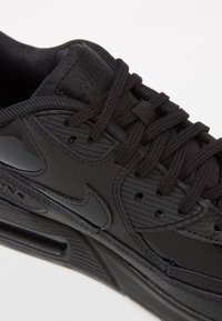 Svart atletisk sko med texturerat läder och syntetiska material, med en vadderad krage, snörningar och glänsande detaljer på sidan.