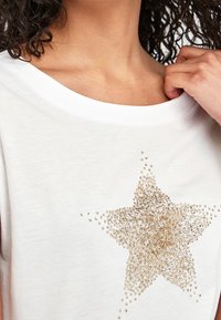 Hvid kortærmet skjorte med et guld palietstjernedesign fortil. Blødt stof med bred halsudskæring og subtil tekstur.