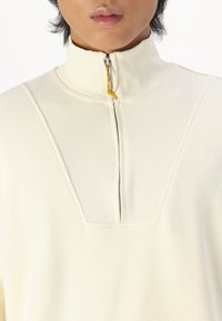 Svijetlosmeđi pulover s visokim ovratnikom, koji ima zatvaranje na pola sa zipom, rebrastu teksturu na ovratniku i glatko tijelo s ušivenim obrubom.