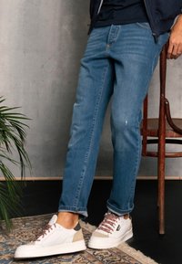 Jeans azul claro de corte recto, combinados con zapatillas blancas que presentan acentos en beige y marrón. El fondo incluye una planta y una silla de madera.