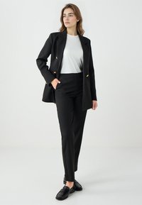 Zwarte op maat gemaakte blazer met gouden knopen, gecombineerd met hoogstaande zwarte broek. Model draagt een witte top en zwarte instappers. Zachte textuur.
