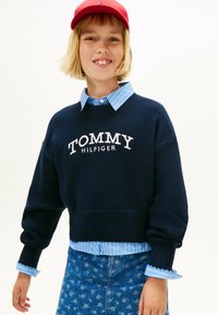 Felpa lavorata a maglia blu navy con logo "TOMMY HILFIGER", abbinata a una camicia color azzurro chiaro a righe con colletto e a un cappellino rosso. Gonna di jeans con motivo floreale.