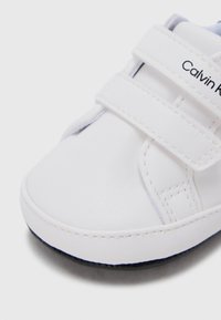 Άσπρο δερμάτινο παπούτσι με δύο λουριά βέλκρο, στρογγυλερή μύτη και αντίθετη σόλα σε ναυτικό μπλε. Διαθέτει το λογότυπο της Calvin Klein στο πλευρό.