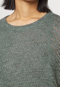 Ljusgrön, stickad tröja med rund halsringning, som har en lös väv och subtil textur, framhävd av öppna stiksaccenter på axlarna.