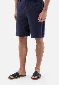 Marineblaue Shorts aus strukturiertem Material mit elastischem Bund. Kombiniert mit schwarzen Rutsch-Sandalen mit schichtigem Design und beigem Sohlen.
