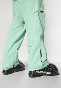 Pantaloni da sci di un verde menta chiaro con tasche con zip e dettagli neri, abbinati a stivali da sci neri con un marchio verde e bianco.