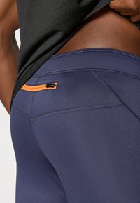 Leggings navy con una textura de tela suave presentan un bolsillo con cremallera naranja en la parte trasera, complementados por líneas de costura y costuras contrastantes.