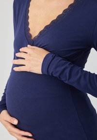 Μακριά μπλούζα εγκυμοσύνης σε σκούρο μπλε ύφασμα, με μακριά μανίκια και βαθύ V-λαιμόκοψη με δαντελένια λεπτομέρεια. Λείος υφασμάτινος χαρακτήρας, εφαρμοστός για να αναδεικνύει την κοιλιά.