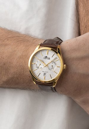 Montre-bracelet avec boîtier en or, cadran blanc texturé, bracelet en cuir marron, sous-cadrans pour le jour et les 24 heures, affichant l'heure à 10h10 et la date au 2, portée au poignet.