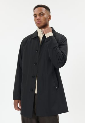 Homme portant un manteau noir boutonné par-dessus un pull beige zippé et un pantalon marron, ajustant son col, debout devant un fond clair uni.