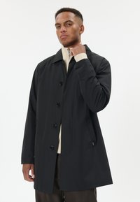 Muž v černém kabátu na knoflíky přes béžový svetr na zip a hnědé kalhoty, upravující si límec, stojící před jednobarevným světlým pozadím.