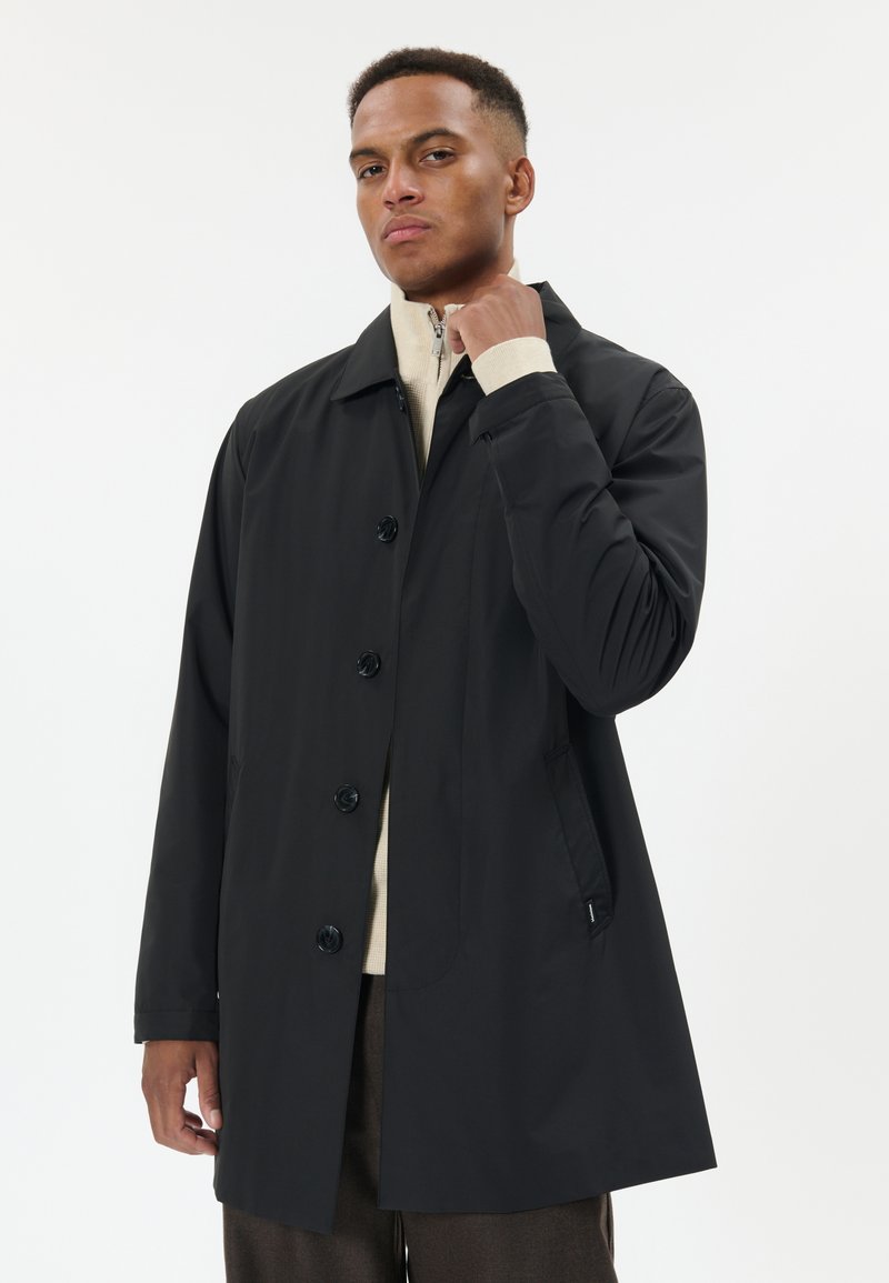 Muž v černém kabátu na knoflíky přes béžový svetr na zip a hnědé kalhoty, upravující si límec, stojící před jednobarevným světlým pozadím.