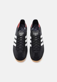 Fekete sportcipő texturált hálós és velúr felsőrésszel, három fehér csíkkal, piros sarokfüllel és gumitalppal.