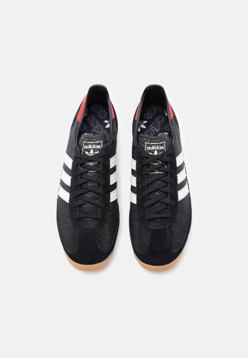 Black athletic shoes with a textured mesh and suede upper, featuring three white stripes, a red heel tab, and a gum sole.