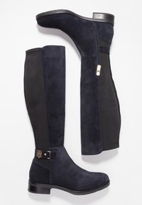 Stivali alti fino al ginocchio in suede blu navy con pannelli posteriori elastici neri, zip dorata laterale e piccola fibbia dorata accentuata vicino alla caviglia.