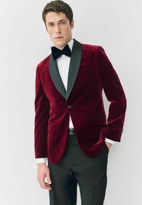 Burgundi bársony zakó fekete selyem sálgallérral, egy gombbal és elülső zsebekkel, fekete nadrággal és csokornyakkendővel párosítva.