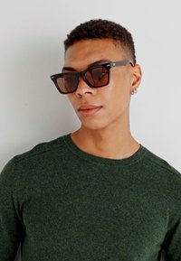 Svarta kantiga solglasögon med brunt tonade linser. Modellen bär en grön texturerad tröja med rundad hals och åtsittande ärmar.