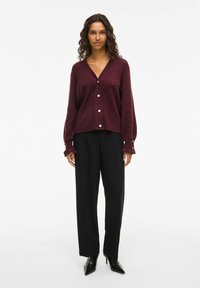 Cardigan in magenta lavorato a maglia con scollo a V, maniche lunghe con polsini a volant e chiusura con bottoni. Abbinato a pantaloni neri a vita alta.