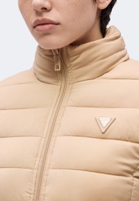 Personne portant une veste matelassée beige avec un col haut et un logo triangulaire Guess sur la poitrine, fermeture éclair partiellement fermée.