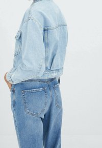 Chaqueta de denim azul claro con diseño recortado, costuras en contraste y un bolsillo en el pecho. Combinada con jeans azul claro a juego.