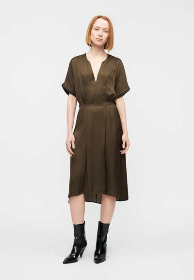 Φόρεμα ελιάς με βαθύ ντεκολτέ σε σχήμα V, κοντά μανίκια και εφαρμοστή μέση, σε συνδυασμό με μαύρα μποτάκια αστραγάλου. Λείο, ελαφρύ ύφασμα.