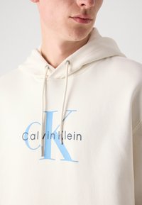 Sudadera beige con capucha ajustable, logo "CK" en azul y texto "Calvin Klein" en gris oscuro. Textura suave con un corte relajado.