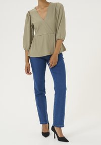 Kvinde iført en lyseolivgrøn wrap-bluse med puffede ærmer, blå straight-leg jeans og sorte spidse højhælede sko, der står foran en ensfarvet baggrund.