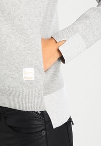 Szara dzianinowa bluza z prążkowanymi mankietami; posiada boczną kieszeń i metkę marki. Tkanina o fakturze; zestawiona z czarnymi dżinsami.