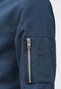 Nahaufnahme eines marineblauen Jackenärmel mit einem vertikalen silbernen Reißverschluss und zwei kleinen rechteckigen Stofftaschen am Oberarm.