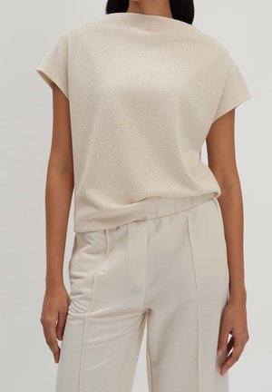 Femme portant un haut texturé crème à manches courtes et un pantalon taille haute crème devant un fond clair.