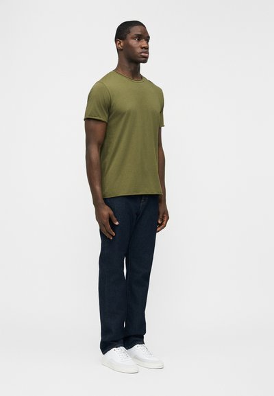 Άνδρας που στέκεται φορώντας μπλουζάκι με κοντά μανίκια σε χακί πράσινο, σκούρο μπλε τζιν και λευκά αθλητικά παπούτσια μπροστά σε απλό φωτεινό φόντο.