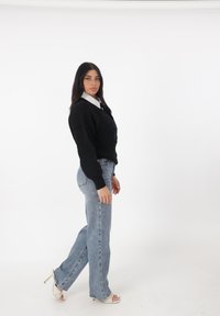 Maglione a costine nero, camicia bianca con colletto sotto, jeans a gamba dritta azzurri chiari e sandali aperti color crema con accenti dorati.