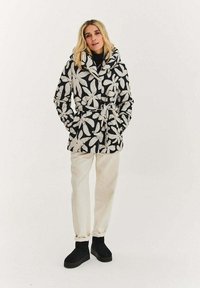 Černobílá bunda s květinovým vzorem, se zavazováním v pase a kapucí, kombinovaná s krémově zbarvenými kalhotami a černými kotníkovými botami.