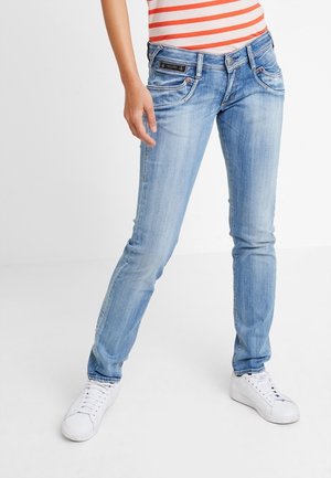 Woman wearing light blue slim-fit jeans, white sneakers, and a red and white striped top, standing with legs apart on a white background.