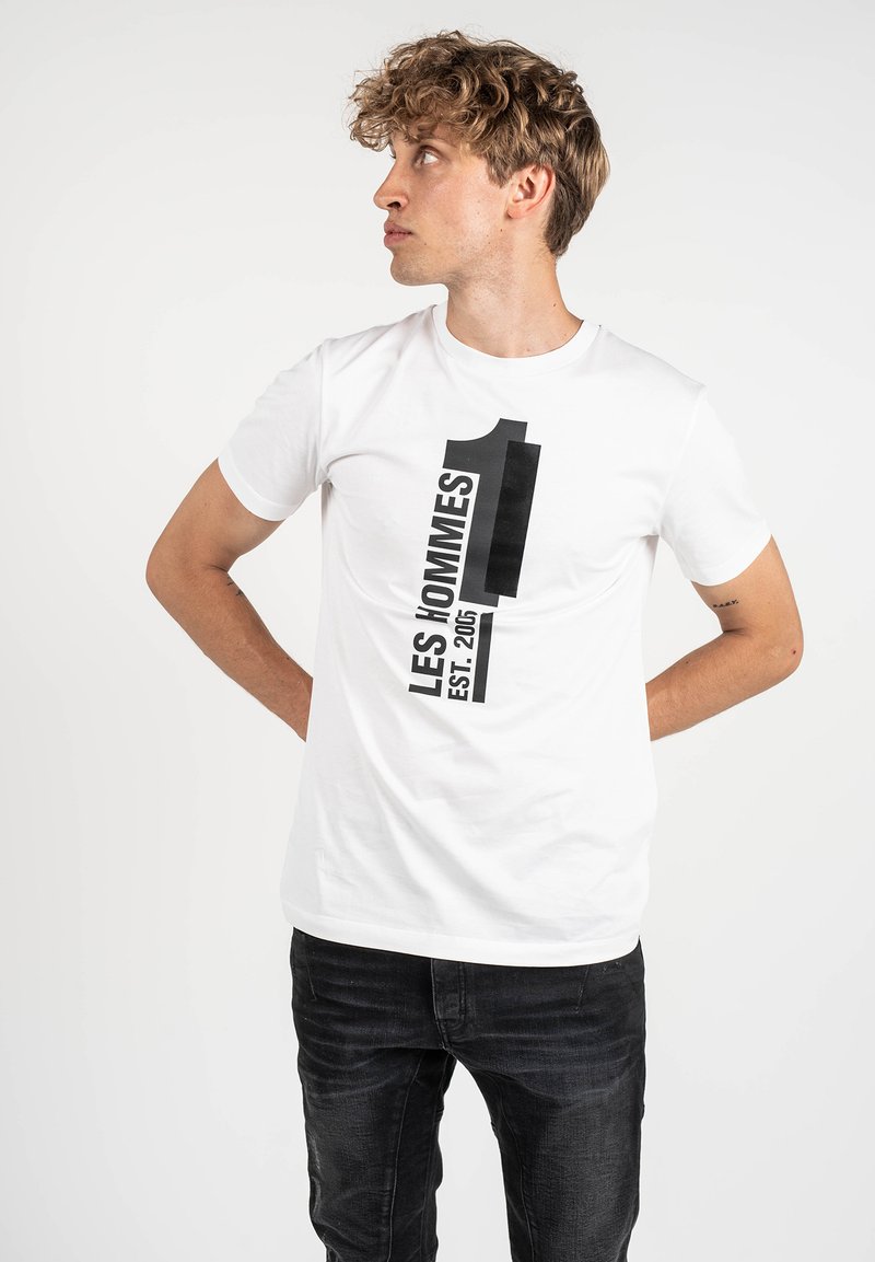 Camiseta blanca de algodón con un estampado gráfico negro que dice "LES HOMMES" y "1 EST. 2005." El modelo lleva jeans negros, con los brazos detrás de la espalda, mirando hacia un lado.