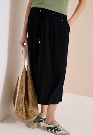 Personne portant une jupe midi noire avec cordon, un haut vert, des baskets beige et vertes, tenant un grand sac fourre-tout tressé avec des anses crème.