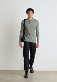 Olijfgroen poloshirt met lange mouwen en een subtiele logo, gecombineerd met zwarte cargo broek en zwarte schoenen, gedragen met een zwarte schoudertas.