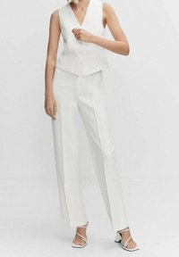 Kobieta ubrana w bezrękawnik z guzikami w kolorze białym, zestawiony z białymi spodniami w wysokim stanem oraz białymi sandałami na obcasie z paskami.