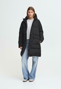 Sort puffermantel med hætte, der har lynlås- og knaplukning, frontlomme og en knælang design. Båret over en lys grå top og jeans.