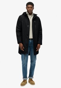 Zwarte waterdichte jas met capuchon, vooraan ritssluiting en zakken, gedragen over een lichtgrijze trui, gecombineerd met blauwe jeans en tan kleurige schoenen.