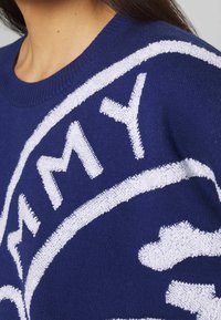 Granatowy sweter z białym teksturowanym logo, okrągłym dekoltem i ściągaczami, prezentujący dzianinę z widocznym detalem ściegów.