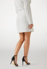 Zwarte sandalen met hoge hakken en een glanzende afwerking, verstelbare enkelband en beige zool; gecombineerd met een jurk met een textuur in wit en zwart patroon.