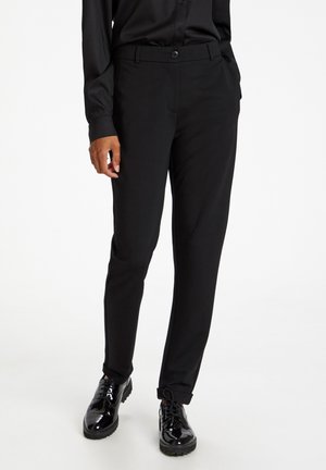 Persoon draagt slim-fit zwarte broek, zwart overhemd met knopen, handen in zakken en glanzende zwarte veterschoenen op een witte achtergrond.