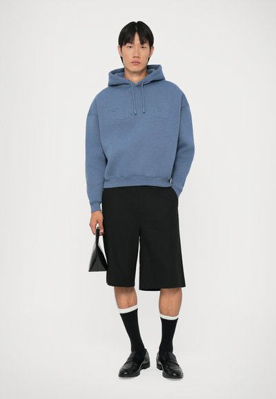 Blauwe sweatshirt met capuchon en reliëflogo, gecombineerd met zwarte shorts, zwarte instappers en zwarte sokken met witte rand, terwijl hij een zwarte tas vasthoudt.
